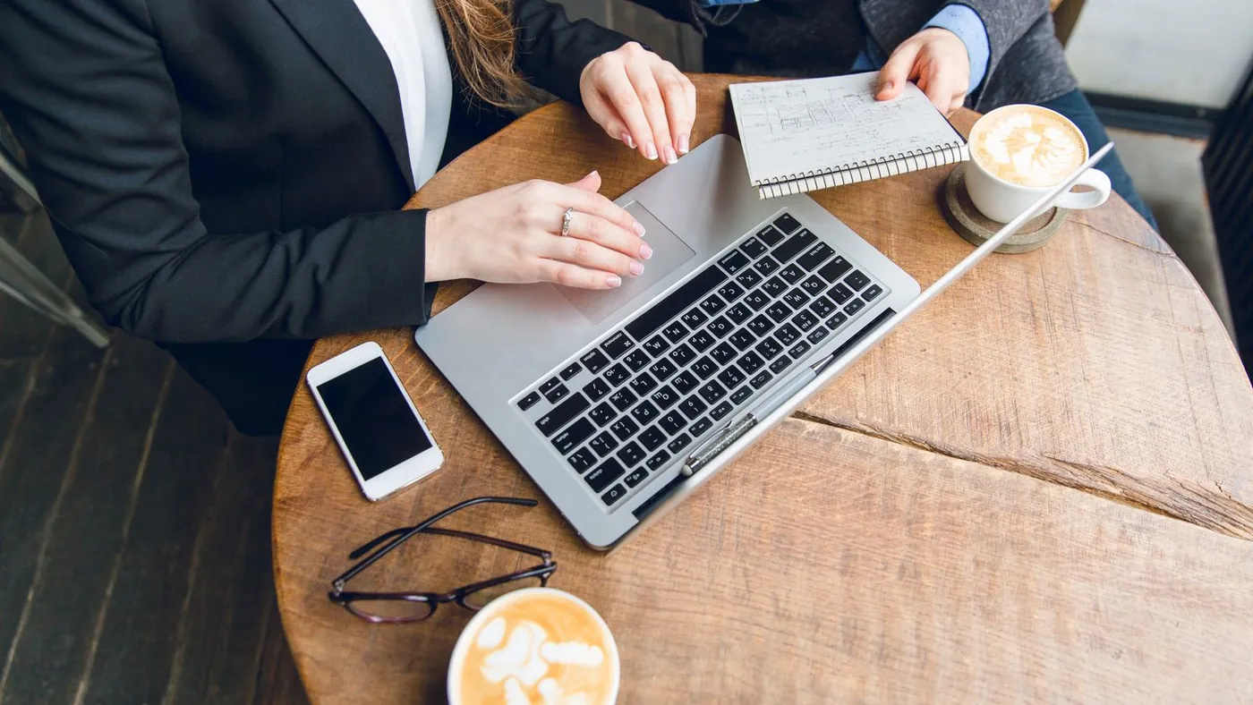 Hands at a laptop with a notebook, phone and coffee on a wooden table
