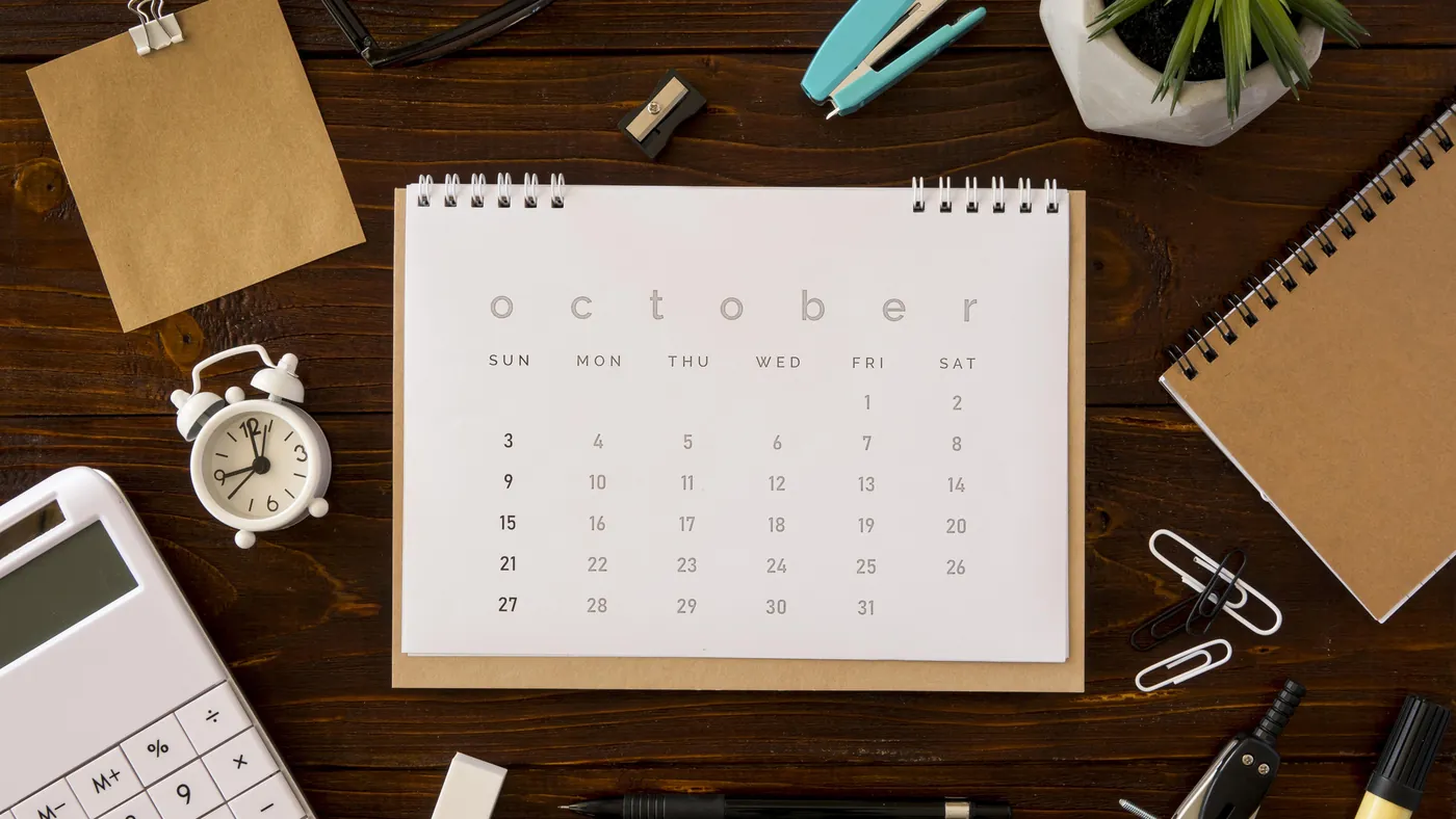 A calendar for October on a wooden table beside a clock and a notebook