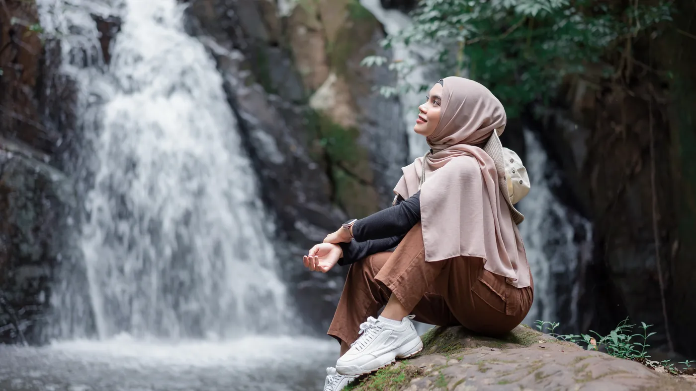 A traveller in a headscarf sitting on a rock beside a waterfall