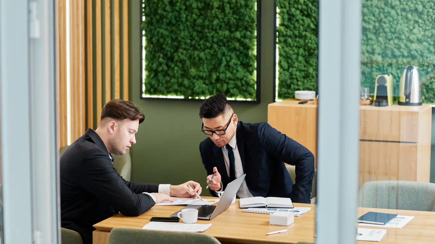 Two people in suits going through papers and a laptop at a meeting table