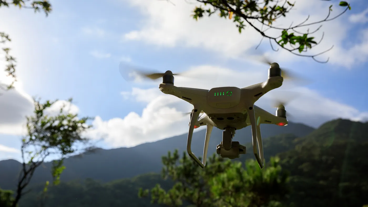 A white camera drone flying above forested hills under a cloudy sky