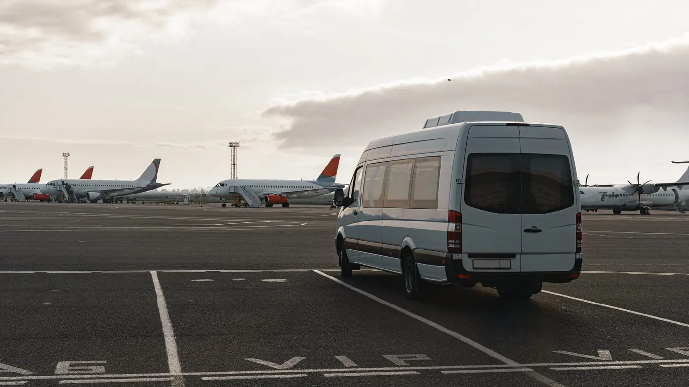 A white minibus parked near an airport apron with aircraft beyond