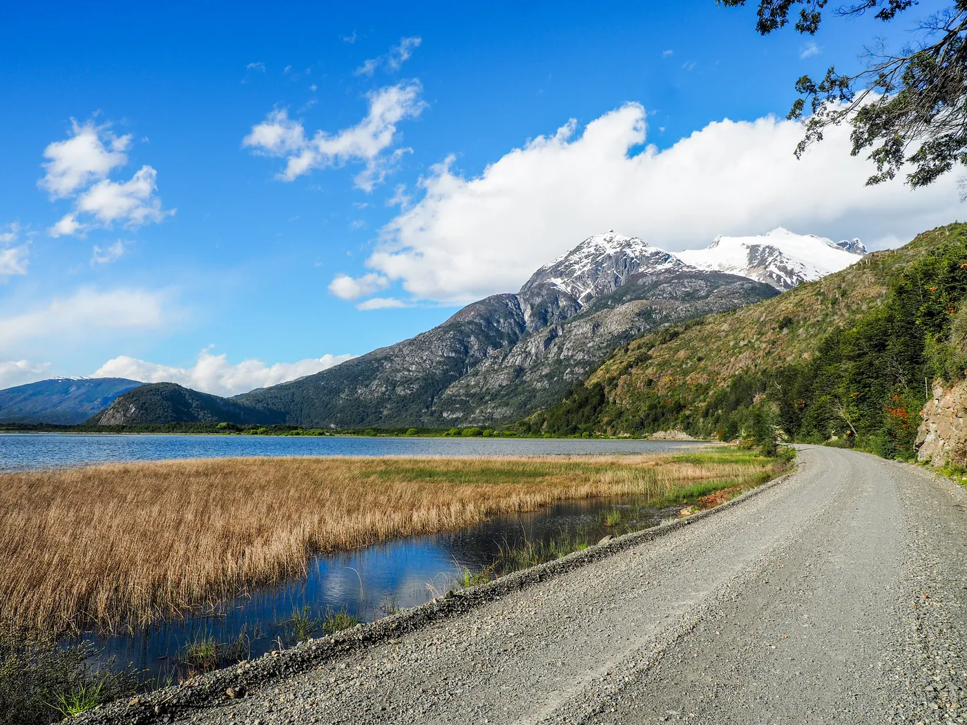A gravel road beside a lake and wetland below snow-capped mountains