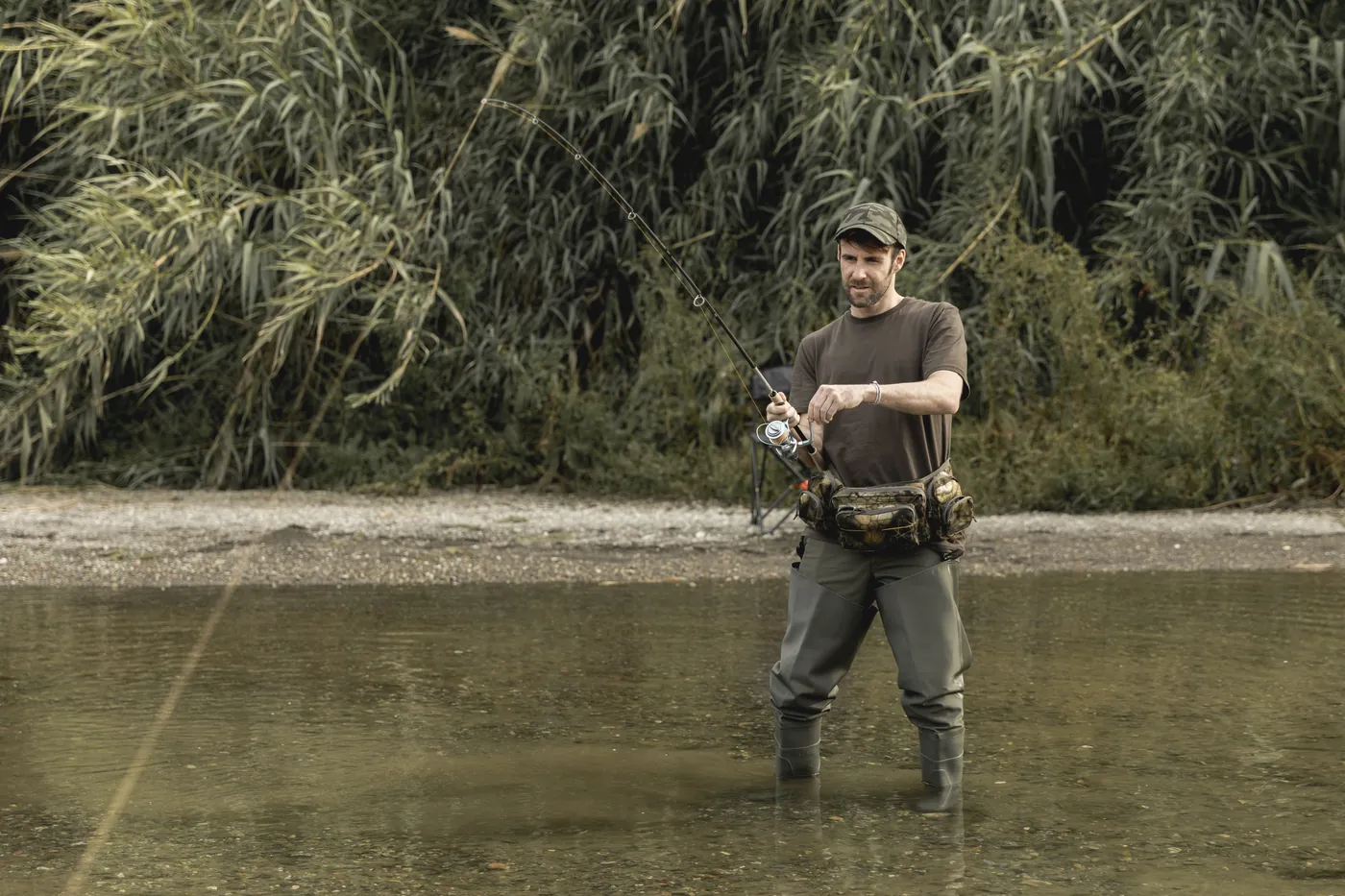 An angler in waders standing in a shallow river with a fishing rod