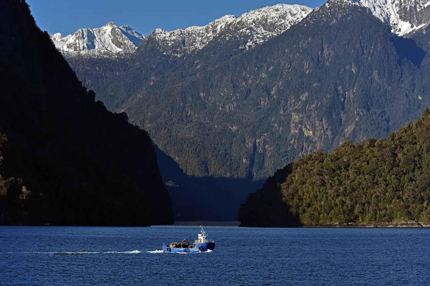 Ein kleines Schiff durchquert einen tiefen Fjord unter schneebedeckten Gipfeln