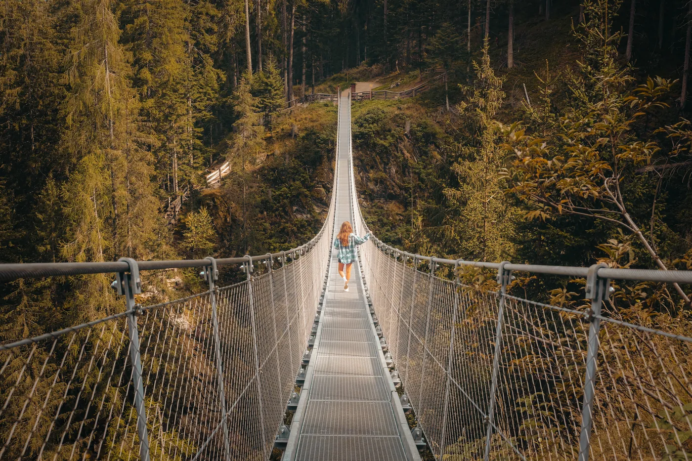 A walker crossing a long suspension footbridge through a forest